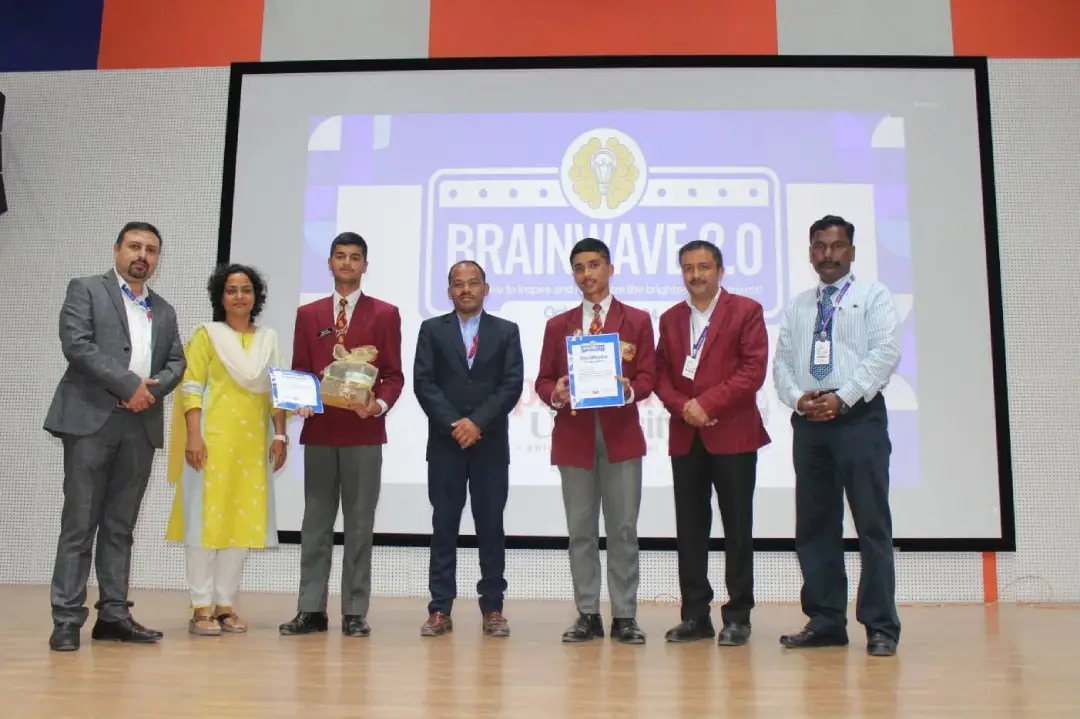 Group of seven people on a stage posing with certificates and a trophy in front of a large screen that says 'BRAINWAVE 2.0'.