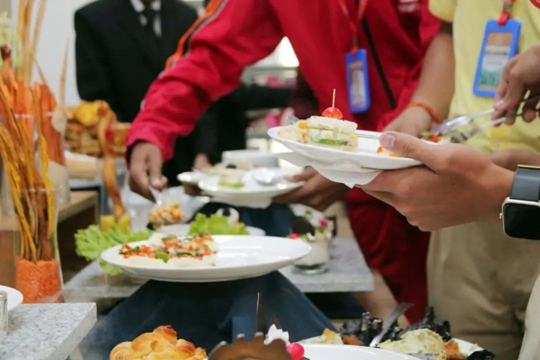 People line up at a buffet, serving themselves plates with assorted appetizers and salads from a long table of dishes.