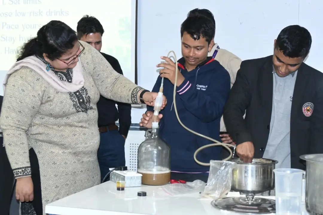 A teacher and three students perform a distillation setup at a lab bench, connecting tubing to a large glass bottle for a chemistry demonstration.