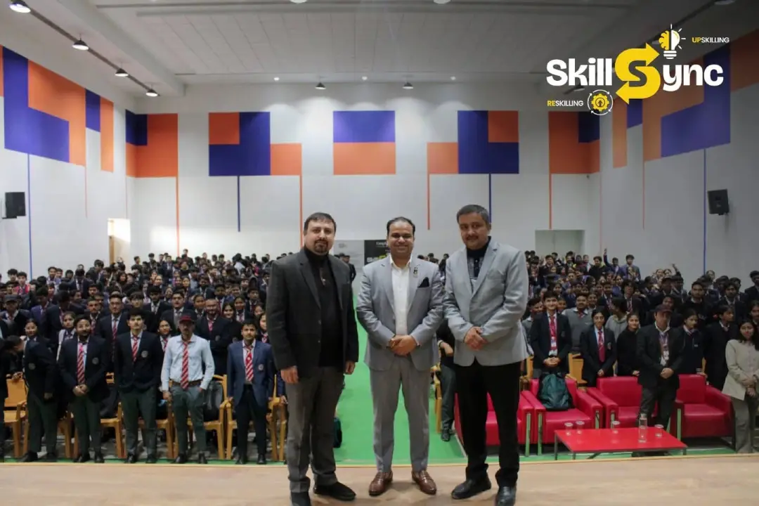 Three men in suits stand on a stage in front of a large audience in a conference hall with colorful wall panels and the SkillSync logo in the corner.