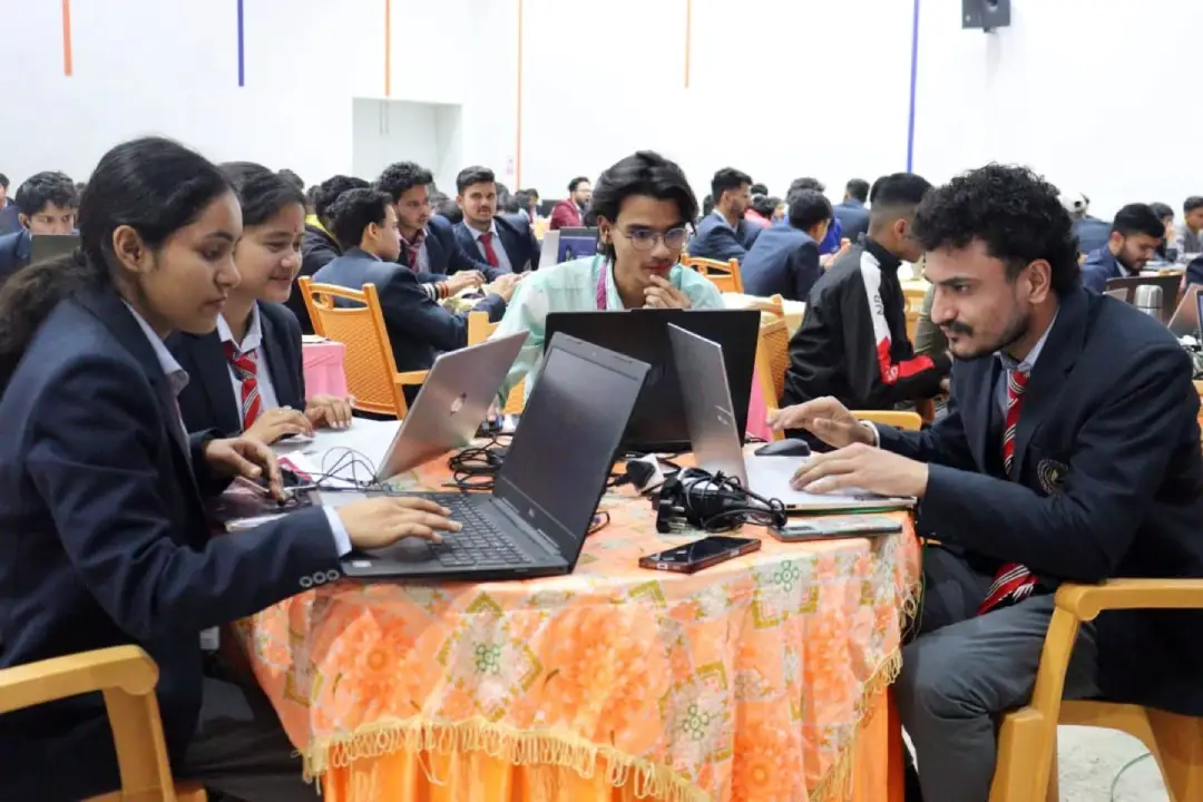 Group of students in suits collaborating around a table with laptops in a busy classroom or lab.