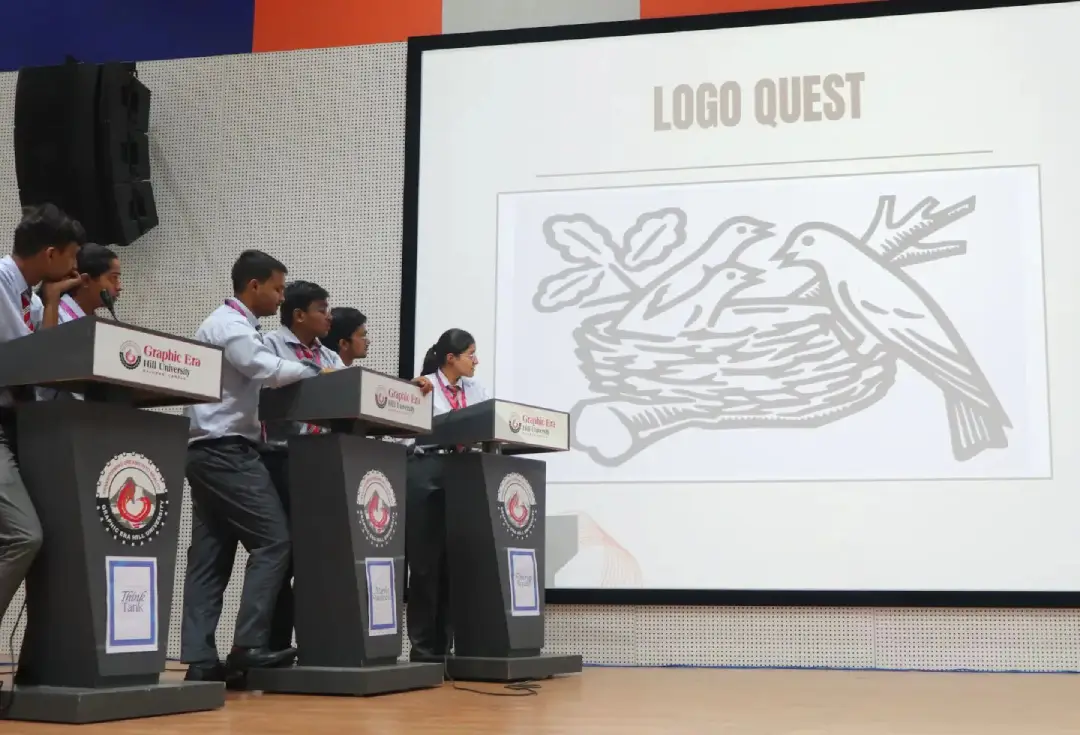 Group of students in uniform at podiums on stage during a Logo Quest event; a screen shows a nest with three birds.