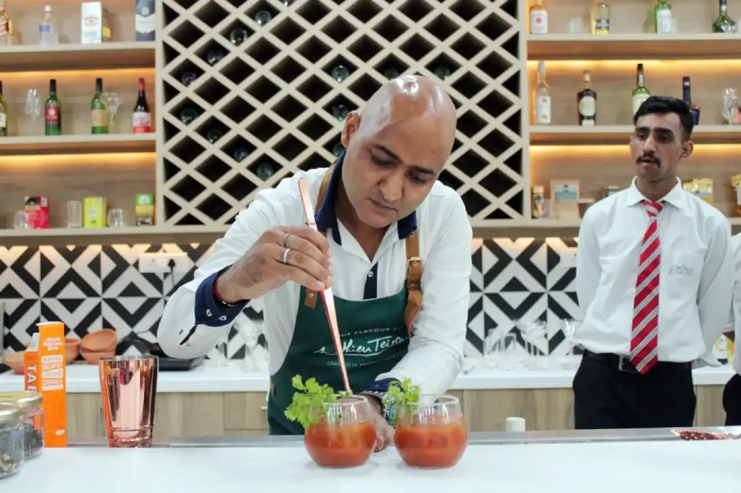 Bartender in a white shirt and green apron garnishes two cocktails with herbs at a modern bar, as a staff member in a striped tie observes in the background.