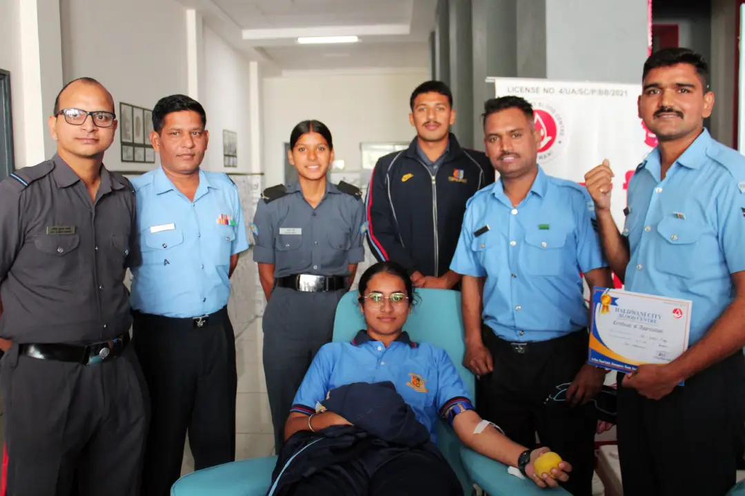 A woman in a blue shirt donates blood, seated in a light blue chair, surrounded by uniformed staff posing for the photo in a clinic. A staff member holds a certificate on the right.