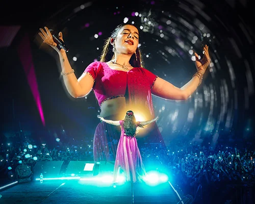 Female singer on stage with arms raised, wearing a red top, bright lights and a cheering crowd in the background.