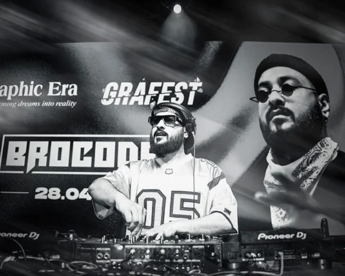 DJ performing at a mixer on stage with a large banner behind him showing 'Graphic Era' and portraits, in black and white.