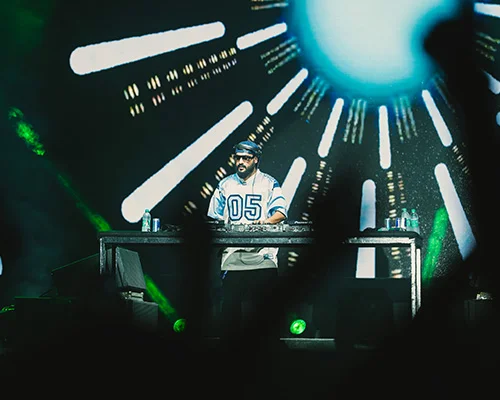 DJ wearing a light-blue jersey with the number 05 performs at a concert, illuminated by a bright blue orb and radial stage lights behind him.