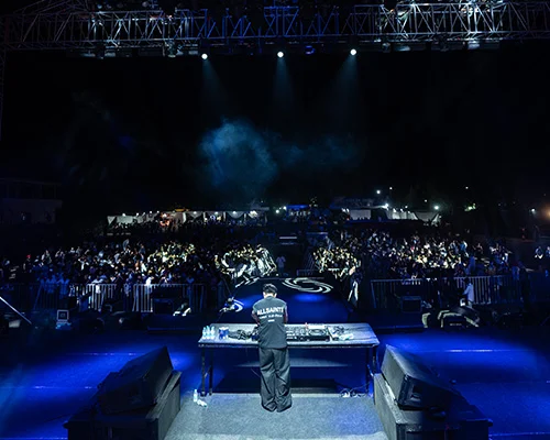 DJ performing on a stage at night with a large crowd in a dimly lit venue, blue lights washing the scene.