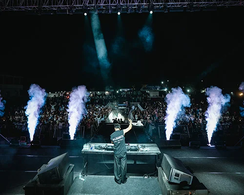 DJ performing on a dark outdoor stage with turntables, raised hands, blue spotlights, and vertical smoke effects as a crowded audience cheers.
