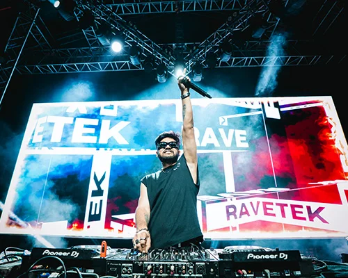 DJ on stage raising a microphone, performing with a mixer in front of a large colorful LED backdrop displaying 'TEK' and 'RAVETEK' amid stage lights and smoke.
