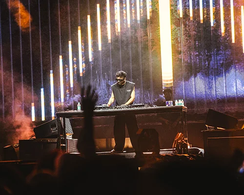DJ in a sleeveless top spins at a desk on a smoky stage with tall colorful LED bars and crowd silhouettes nearby