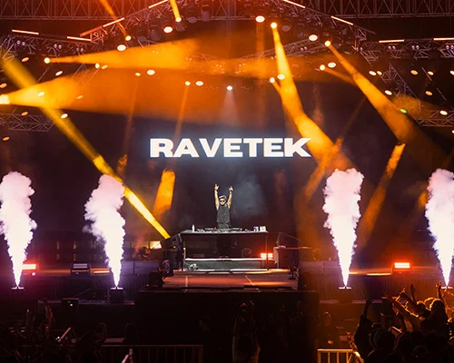 DJ performing on stage with yellow spotlights and white smoke, backdrop shows the word 'RAVETEK' with crowd in the foreground.