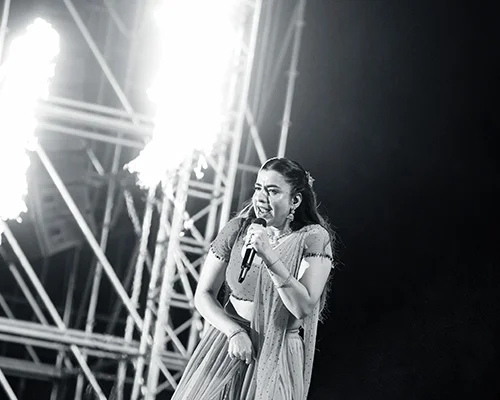 Female singer performs on a lit stage, holding a microphone with metal truss structures in the background.