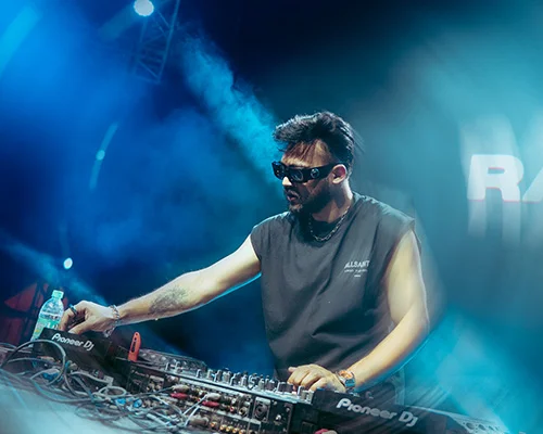 DJ wearing sunglasses and sleeveless shirt performs at a club, hand on mixing console with Pioneer DJ gear amid blue stage lights and haze