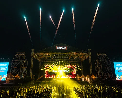 Nighttime outdoor concert with a large stage, yellow spotlights on the crowd, and fireworks overhead.