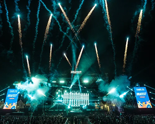 Outdoor night concert with bright fireworks above a large stage and cheering crowd.