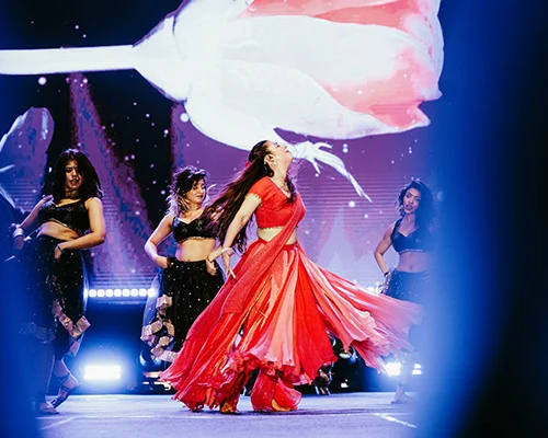 A woman in a flowing red dress spins center stage, with backup dancers in black nearby under bright stage lights.