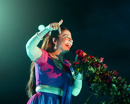 Female singer on stage holding a bouquet of red roses, smiling and pointing upward with one hand.