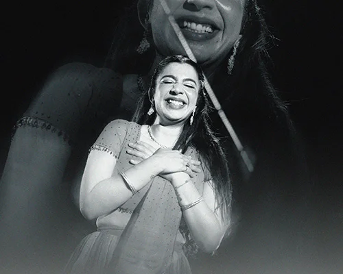 Joyful woman in a formal gown on stage, arms crossed over her chest while smiling; a large, faded portrait of her appears behind and above, with fog at the bottom edge.