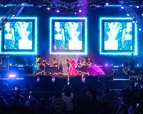 Live concert on a lit stage with dancers and a singer in pink, three large video screens behind them.