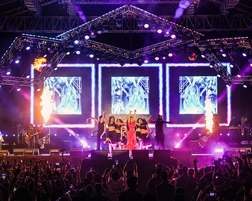 Live concert on a lit stage with dancers, performers, and large screens, crowd cheering in front.