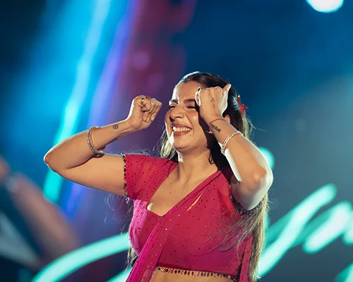 Woman on stage dancing and smiling, wearing a pink outfit with jewelry, raised fists in a celebratory pose.