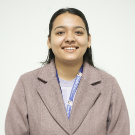 megha-joshi-management-hld Portrait of a smiling woman with dark hair wearing a mauve blazer and a blue lanyard, standing against a white background.