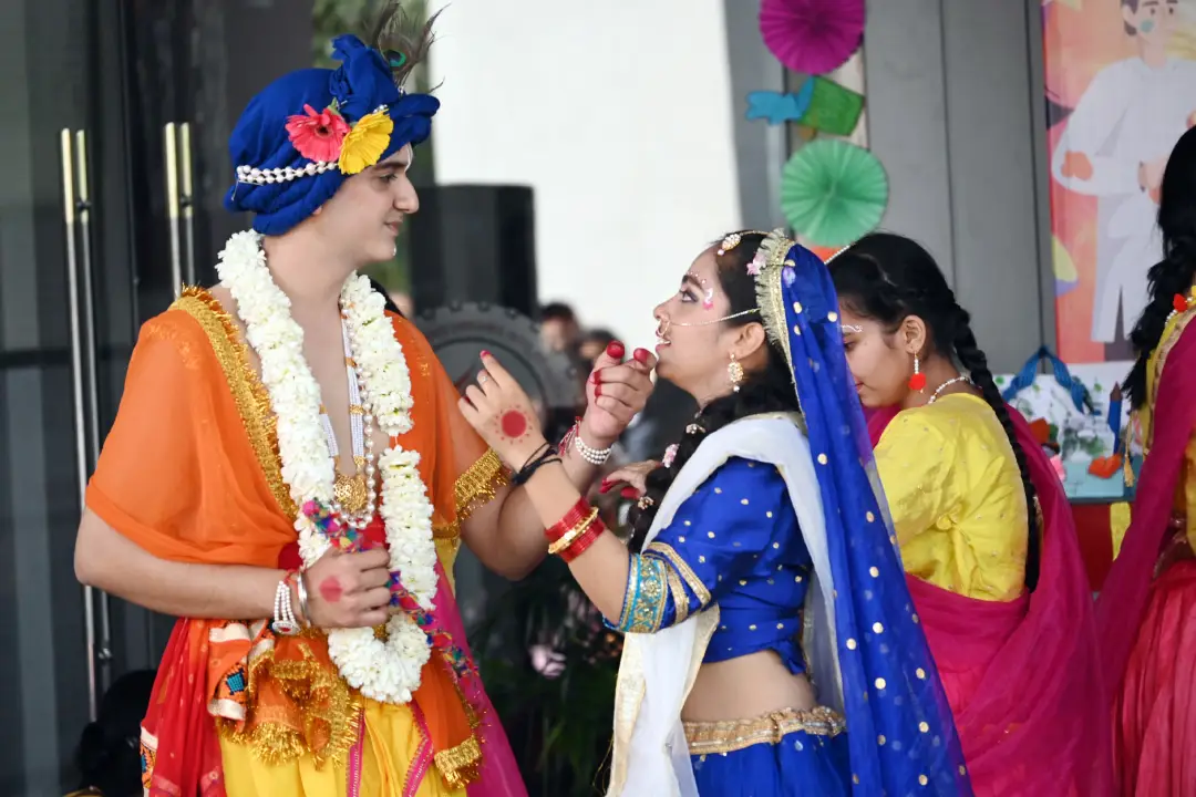 Man in bright orange traditional dress and blue turban with flower garland receives a sweet from a woman in a blue and white lehenga during an Indian wedding ceremony, with colorful decorations in the background.
